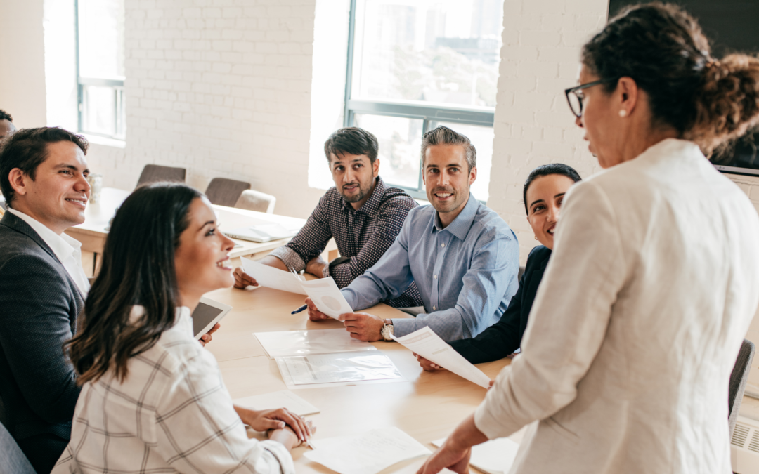 Photo of a team sitting around a table looking at a woman standing who is talking.